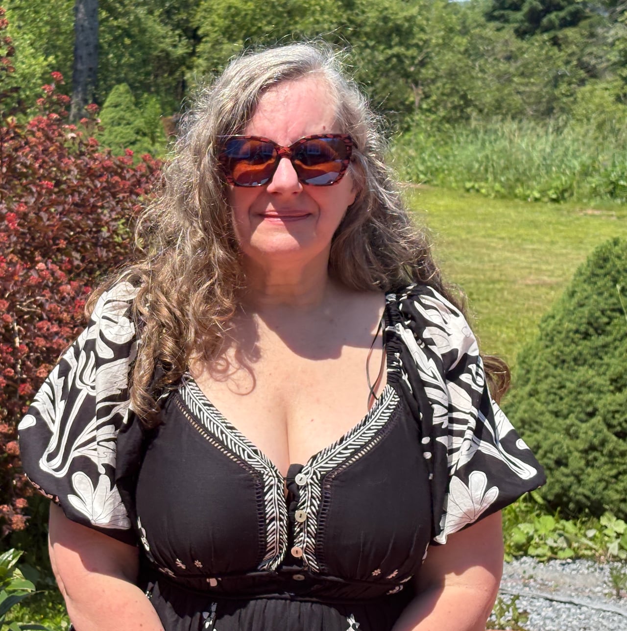 Woman, wearing dress and sunglasses, smiling at camera on a sunny day.