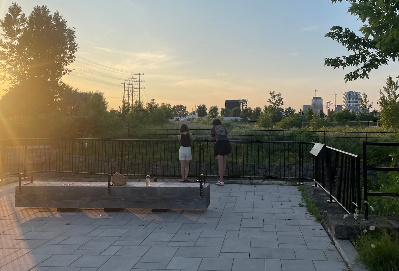 Two people lean on a fence near a path, listening to music coming from a festival across a field.