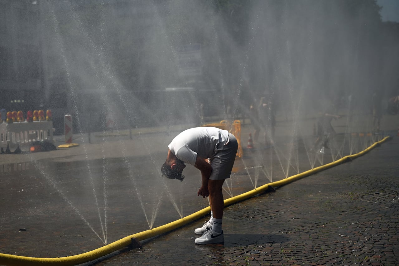 A man, wearing a white t-shirt, grey shorts and a white ball cap, cools off on a hot day by standing bent over a long yellow hose with water spraying out of it.