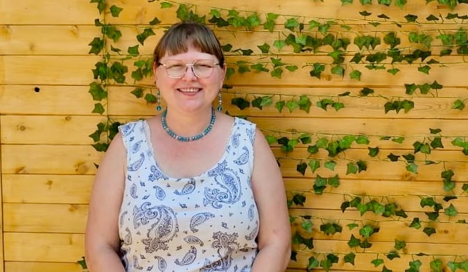 A woman with brown hair pulled back and glasses, smiles at the camera while standing in front of a wooden fence with a vine in summer.