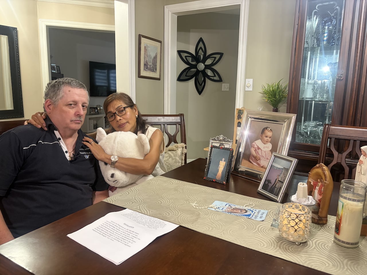 A man and woman sit at a dining table surrounded by photos of their daughter.
