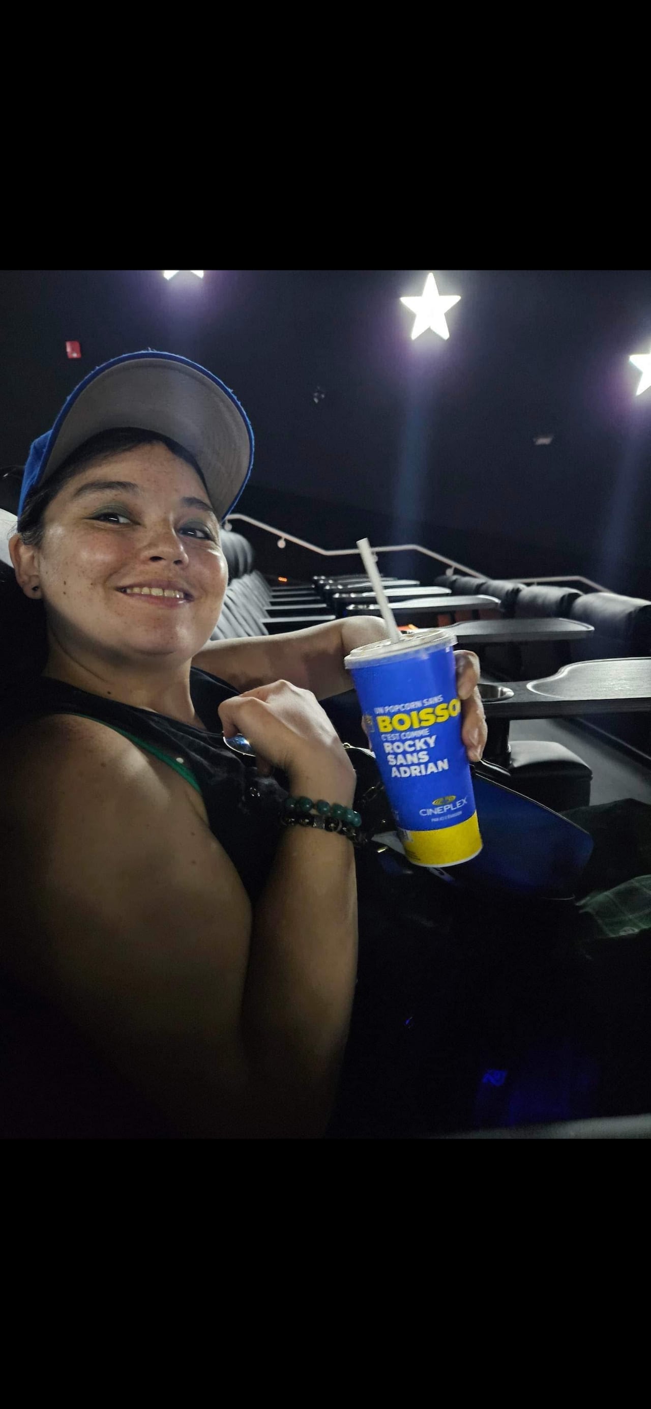A woman sits in a movie theatre seat, smiling at the camera and holding a drink.