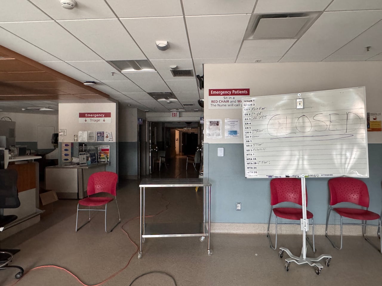 Empty chairs sit in a room with a dark hallway beyond. A whiteboard says "CLOSED." There is a counter and health-care equipment to the left of the picture.