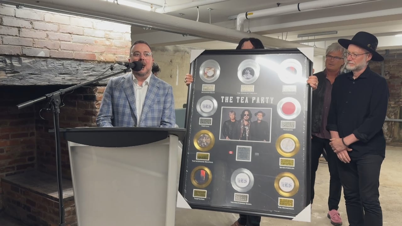 Band members hold an enormous plaque with multiple gold and platinum albums on it while a member of their management team speaks into a microphone at a podium.