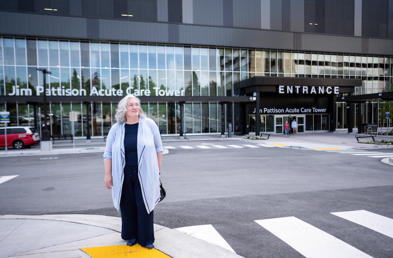 A woman in a long grey cardigan and black clothes stands in front of a hospital building.