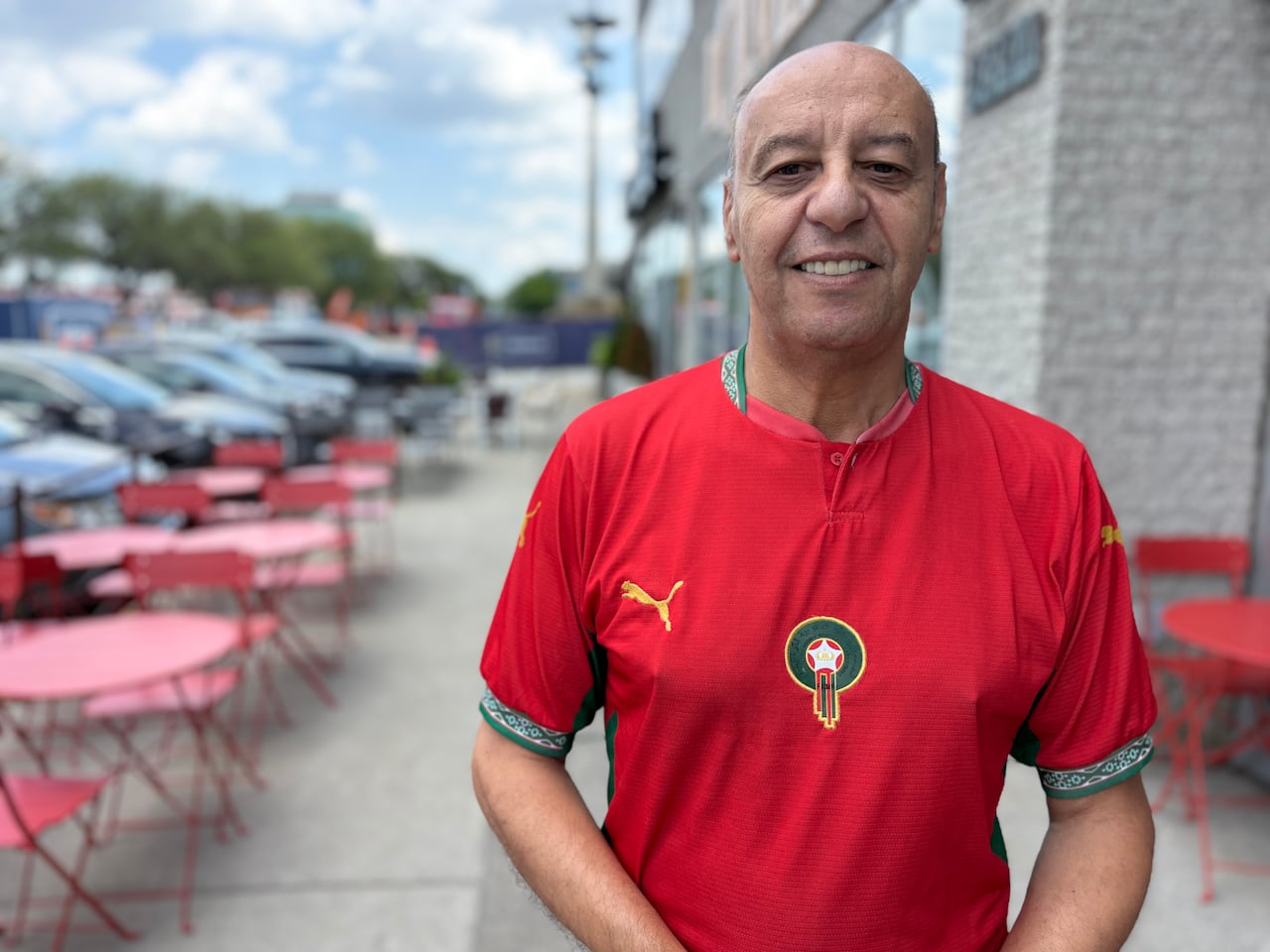 A man stands on the sidewalk amid chairs and tables at a cafe.