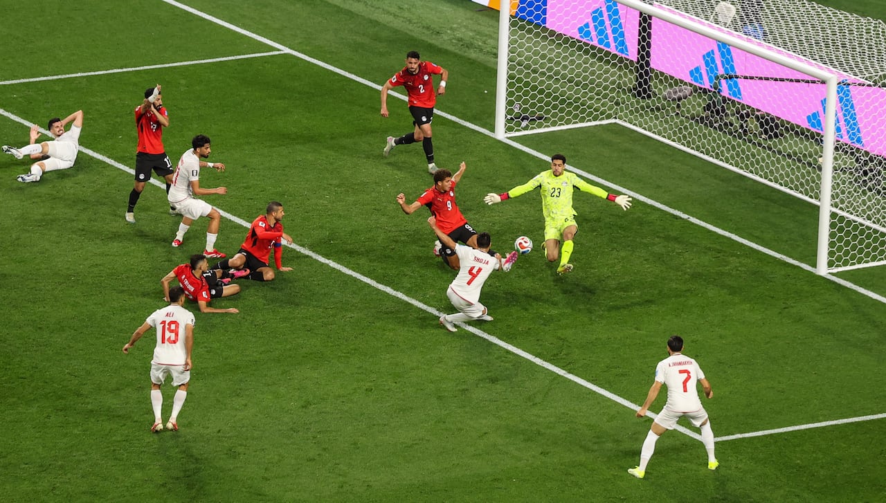 A group of soccer players in either red shirts or white shirts, with one white-shirted player shooting the ball past a goalkeeper in yellow.