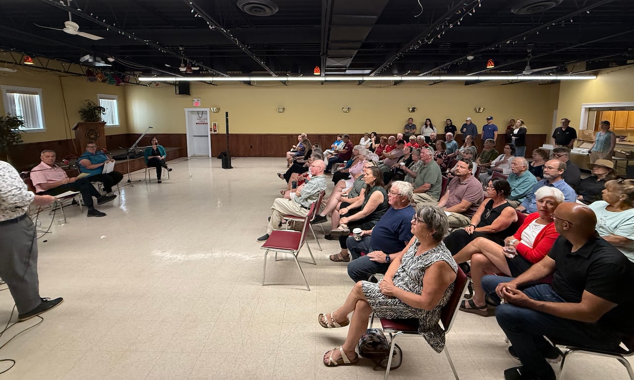 A wide-angle view of an audience seated in rows of chairs listening to three speakers paneling a discussion in a large community room.