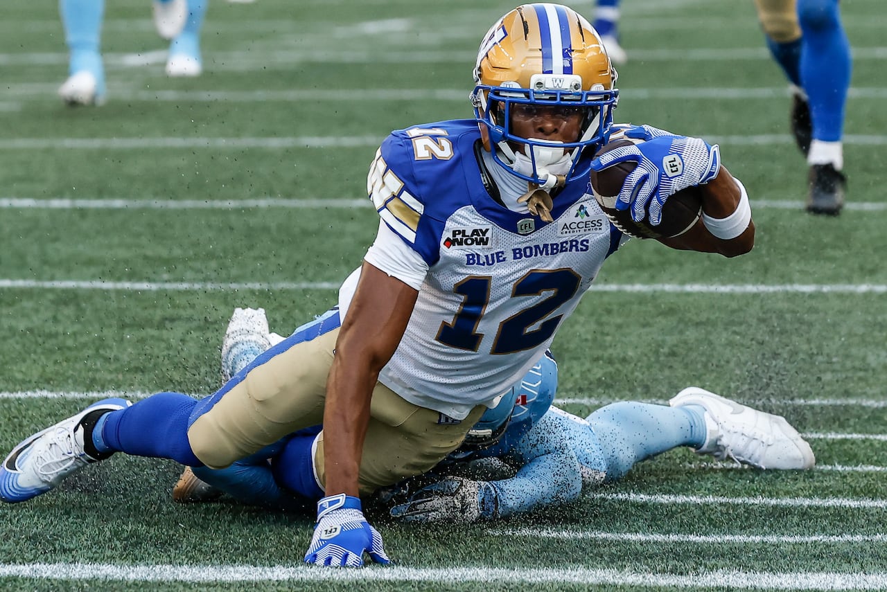 A football player in a blue white and gold uniform dives across a line painted on the green turf as another player in light blue is behind him