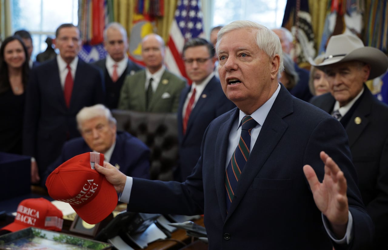 A man with white hair, wearing a navy suit and tie and holding a red ballcap in his right hand, gestures while speaking before a group of people in an ornate office.