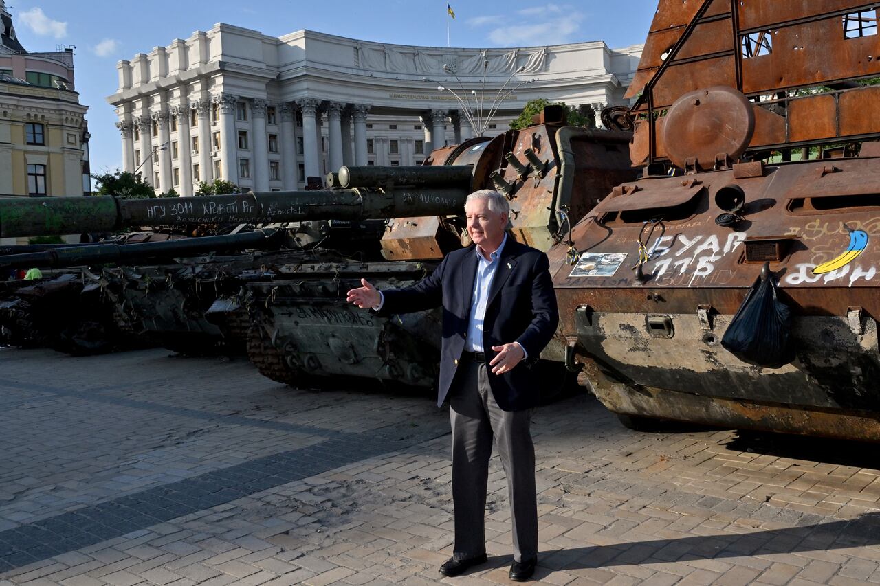 A man in a navy blazer gestures with his right hand as he stands in an outdoor square, with tanks and other military equipment behind him.
