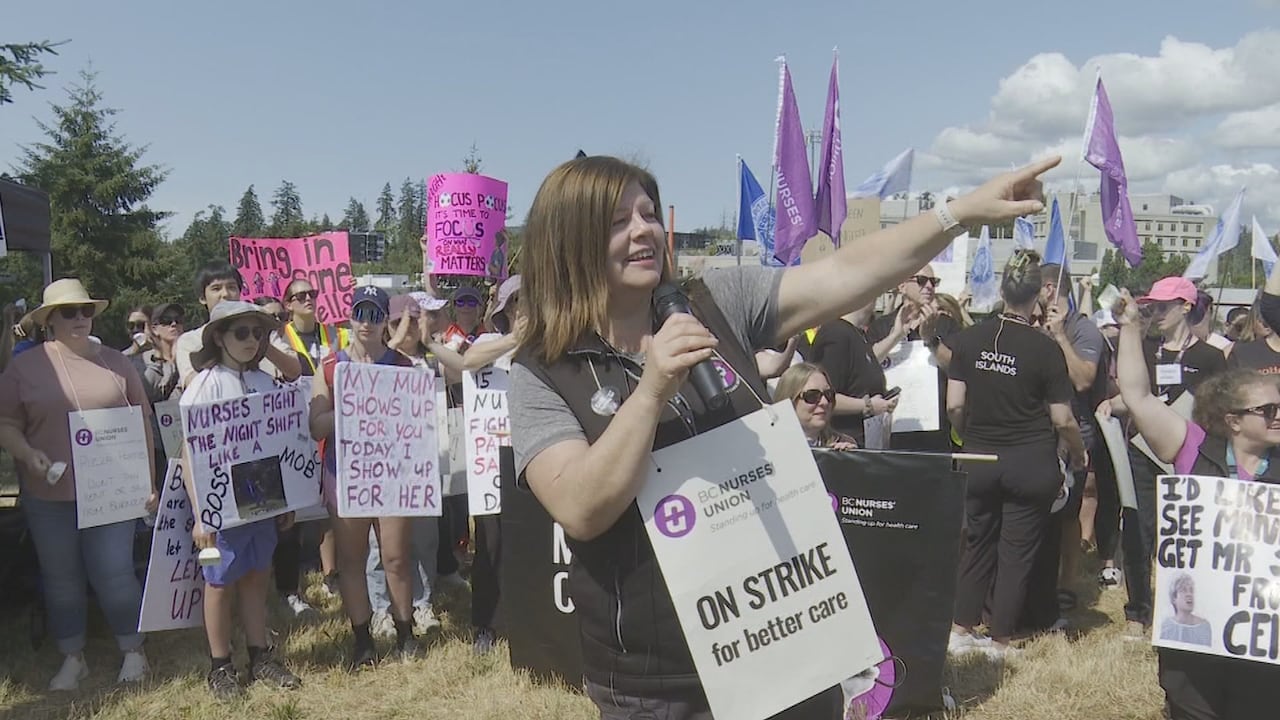 A woman with long brown hair holds a microphone and points at a crowd. She is wearing a picket sign that says "On strike for better care."