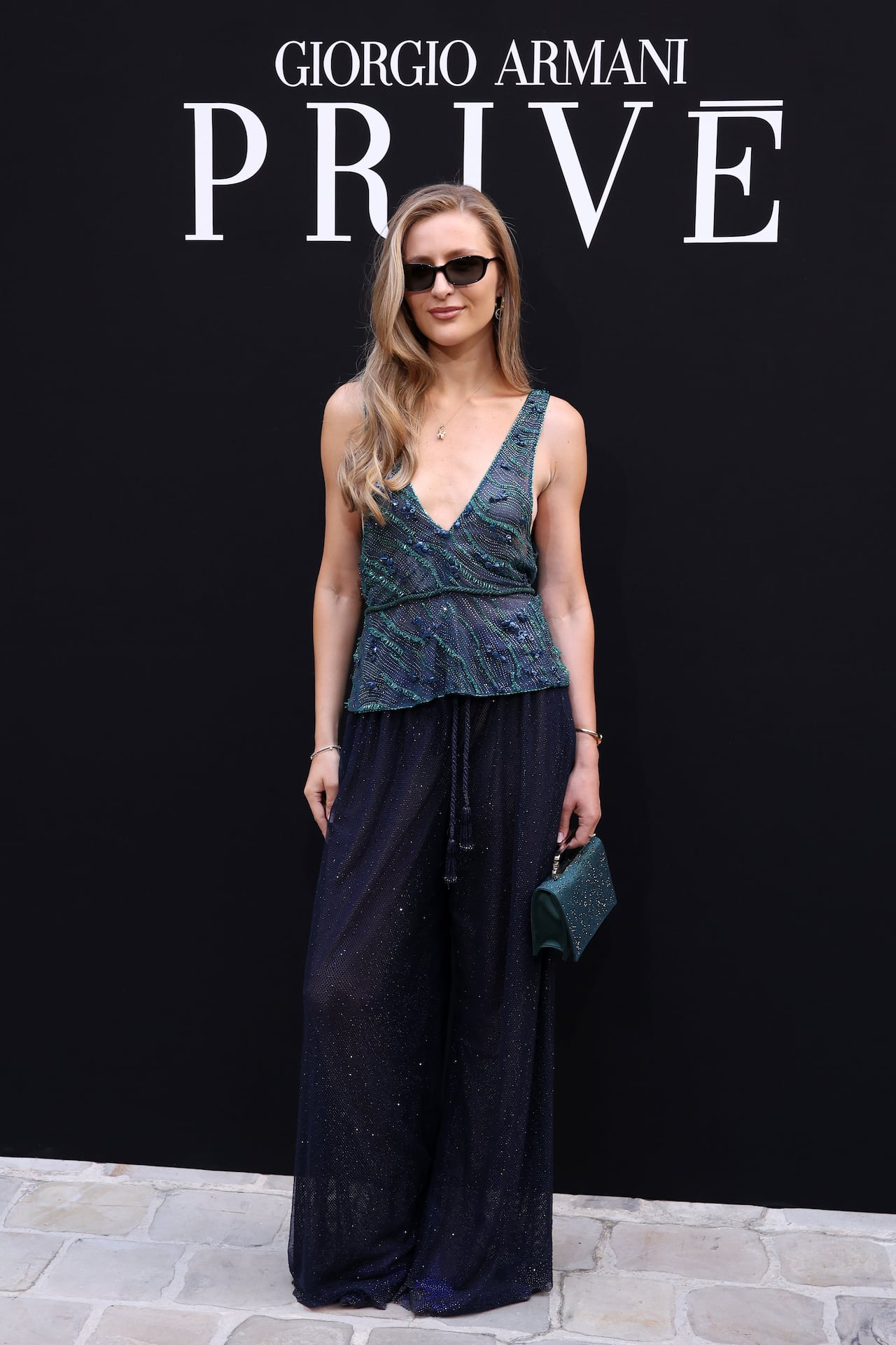 A woman in sunglasses and flowy pants poses in front of a backdrop that says Armani