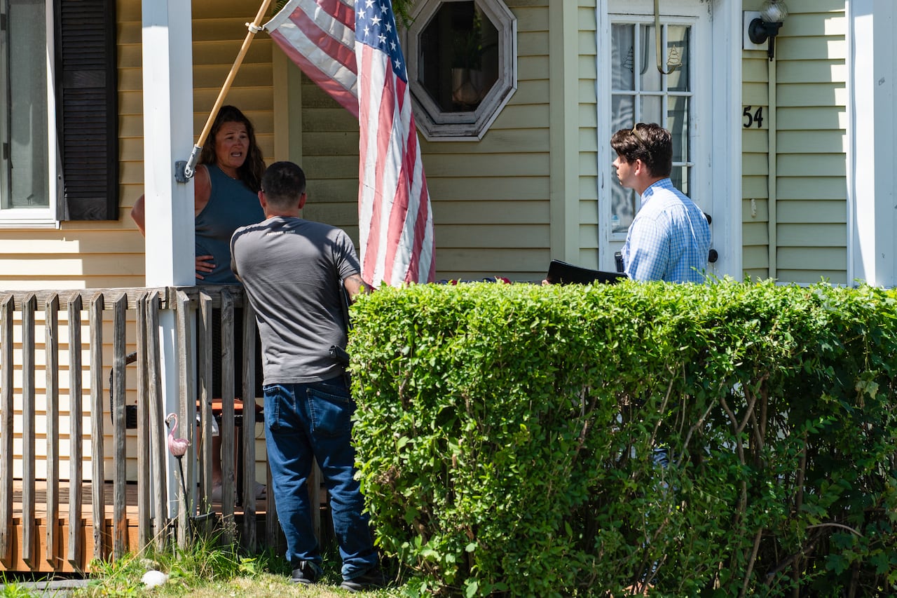 A woman standing on her veranda, with two FBI investigators standing near a hedge while speaking to her.