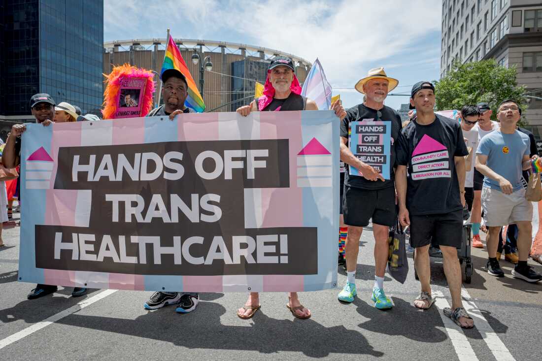 Several people carry a large banner that says "Hands off trans healthcare" as they walk down a city street. Another person has a small sign that says the same thing.