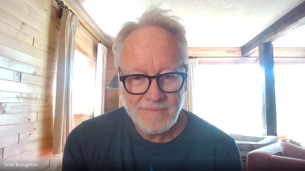 A man with grey hair and glasses sits in a log cabin home.