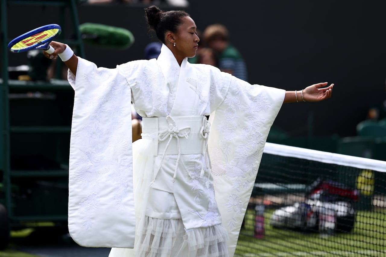 A woman stands on the tennis court holding a tennis racket and wearing a white embroidered kimono-style robe over a tulle skirt.
