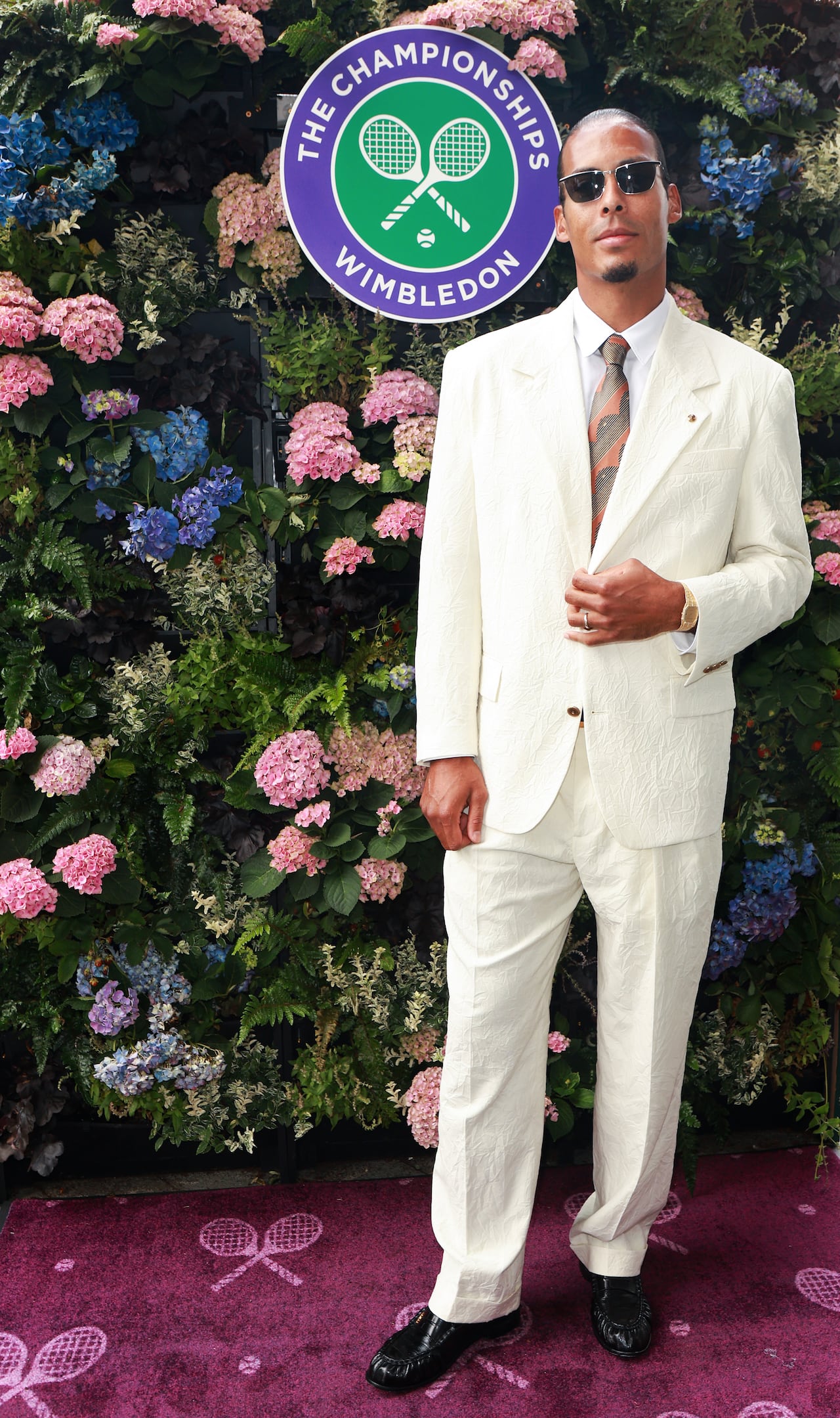 A man poses for a photo wearing a cream suit and sunglasses.