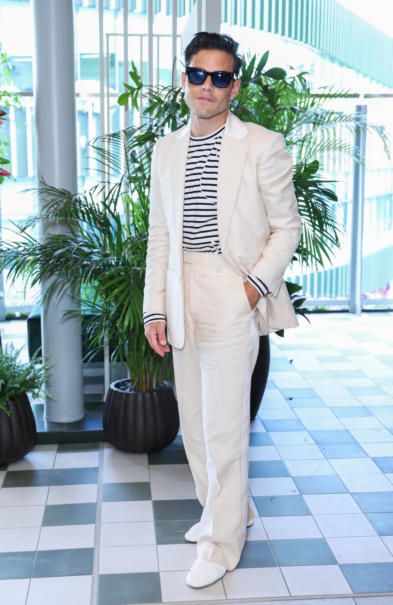 A man poses for a photo wearing a cream-coloured suit with a black and white striped long-sleeve T-shirt and sunglasses.