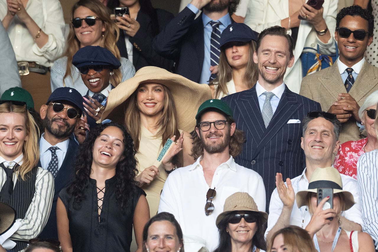 A man and woman sit, watching tennis amongst a crowd. She's wearing a large brimmed hat and beige dress. He's wearing a navy pin-stripe suit.