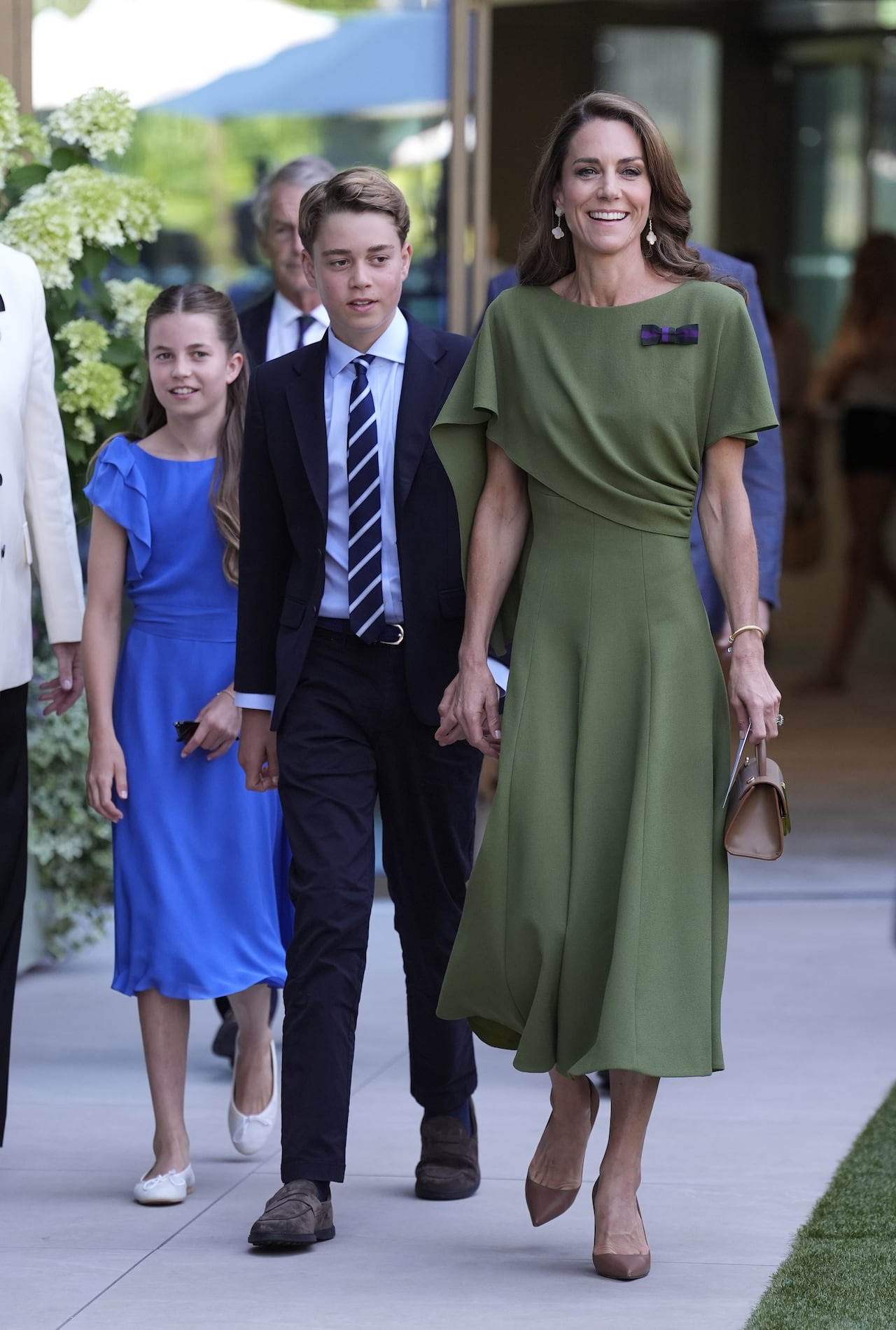 A woman and two kids walking. She's wearing a green dress. The son is wearing a navy suit and the daughter is wearing a blue dress.