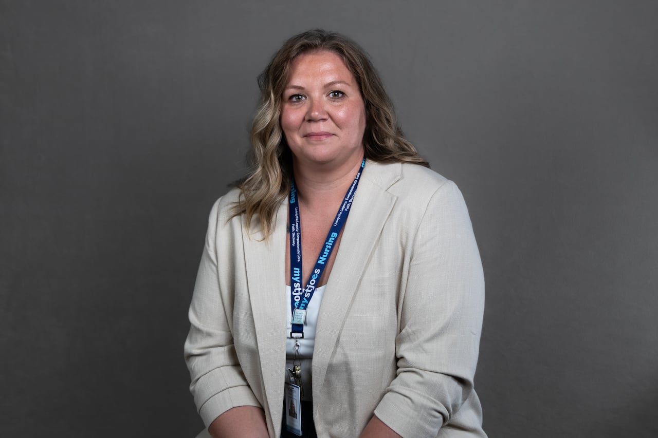 A woman in a beige blazer sits in front of a grey background.