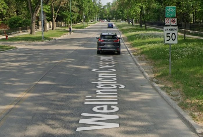 A car drives along a treed street with a bike route sign and speed limit posted on the boulevard.