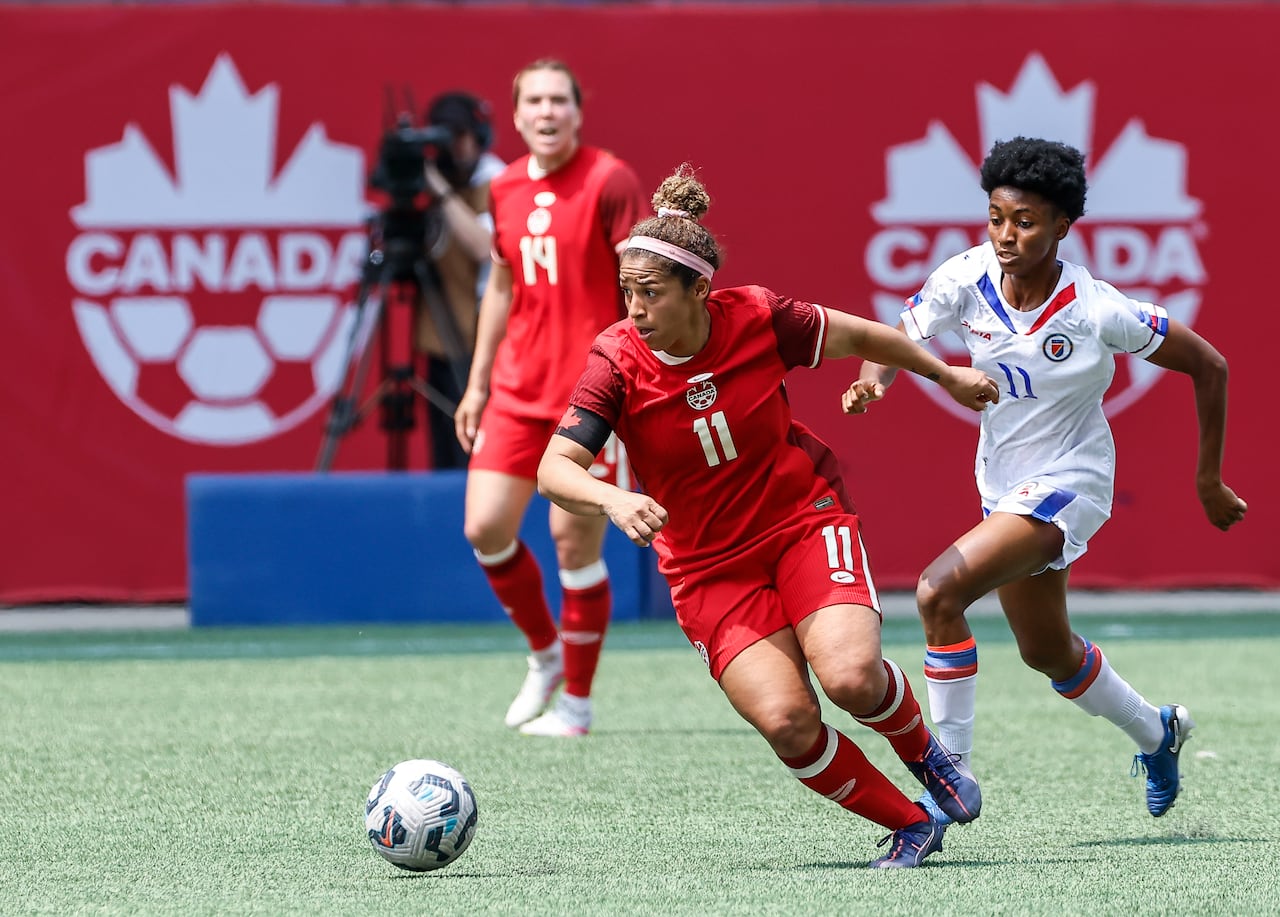 A woman in a red soccer uniform moves toward a soccer ball, with a woman in white in pursuit. The wall behind them has a Canada soccer logo.