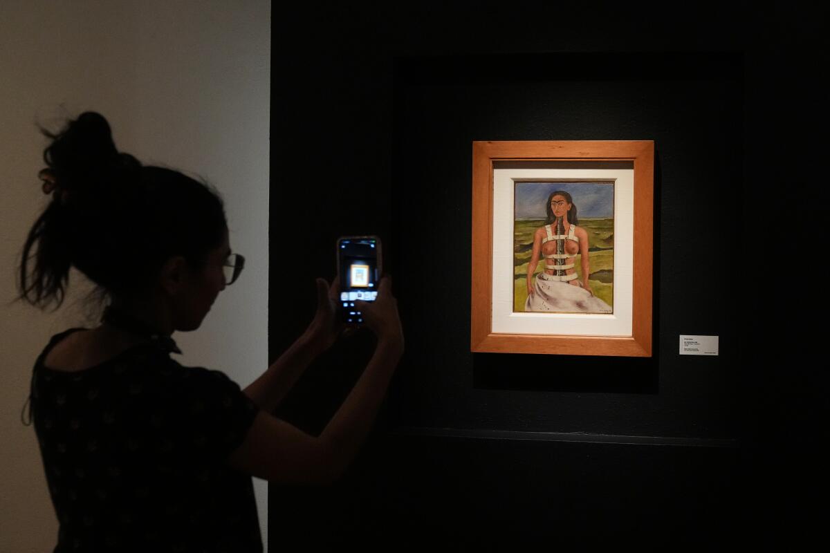 A person uses their cellphone to photograph a small painting on a wall.