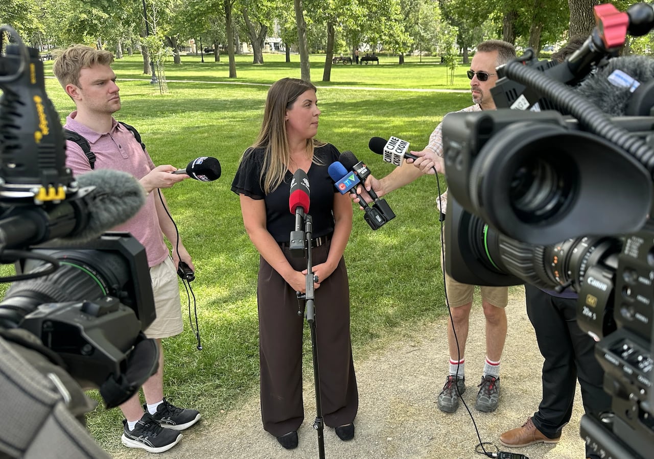 A woman wearing a black shirt and dark pants stands surrounded by reporters in a park setting.