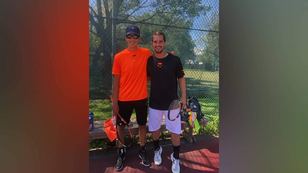 Two boys stand beside each other holding tennis rackets.