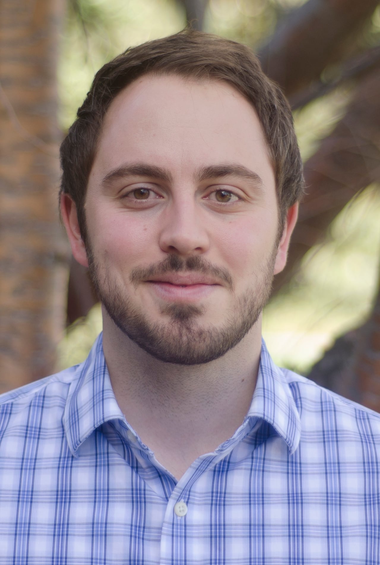 A white man with brown eyes, a brown beard, and brown hair smirks at the camera while wearing a blue and white plaid shirt.