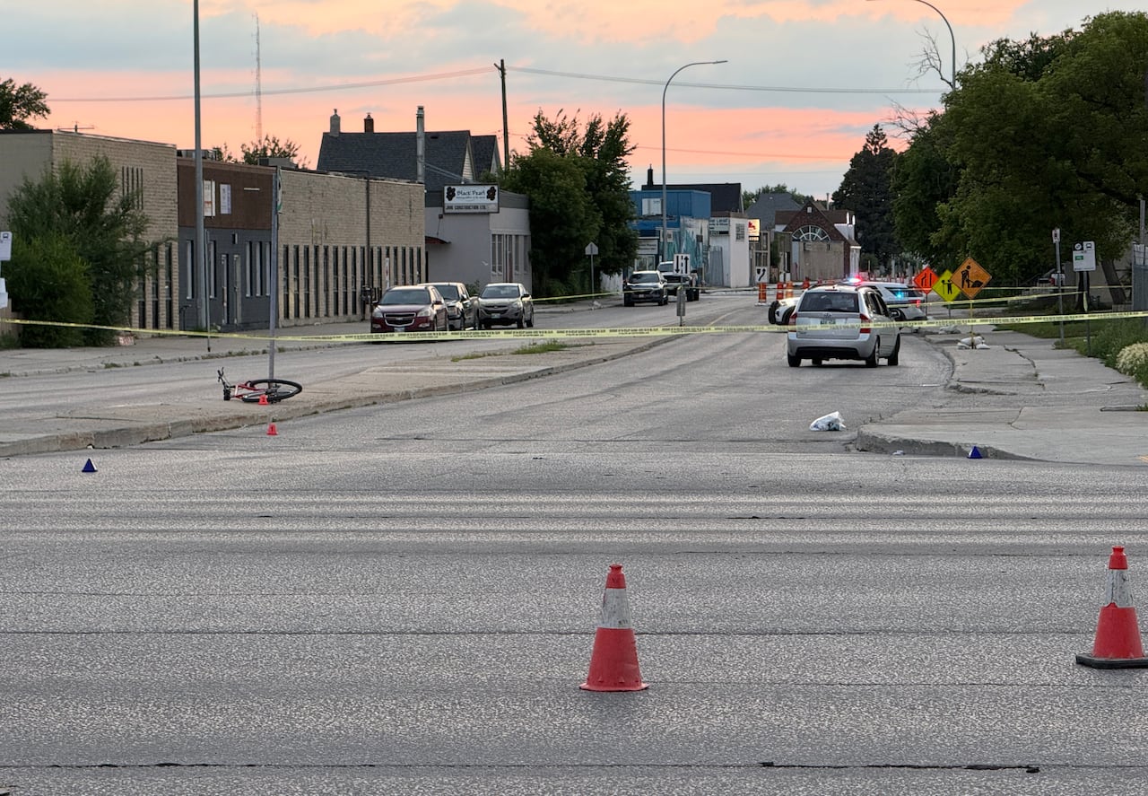 A bicycle lies in the background on a road median. Yellow police tape and orange pylons mark an investigation scene.