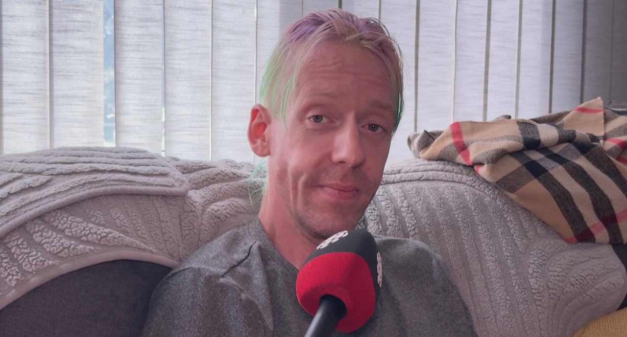 A man sits with a microphone in front of him, looking into the camera