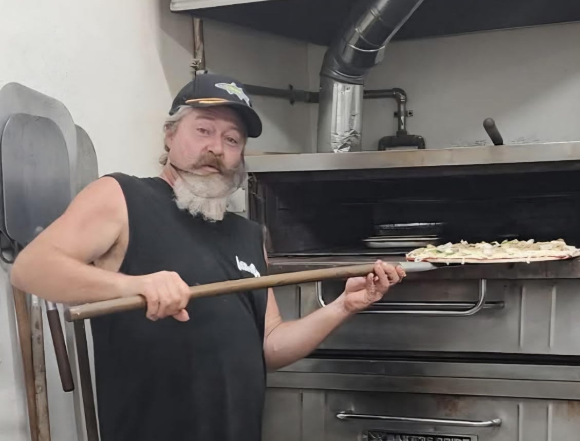 A man with a long gray beard, wearing a black tank top and shorts, poses while he puts a large pizza into a massive pizza oven.
