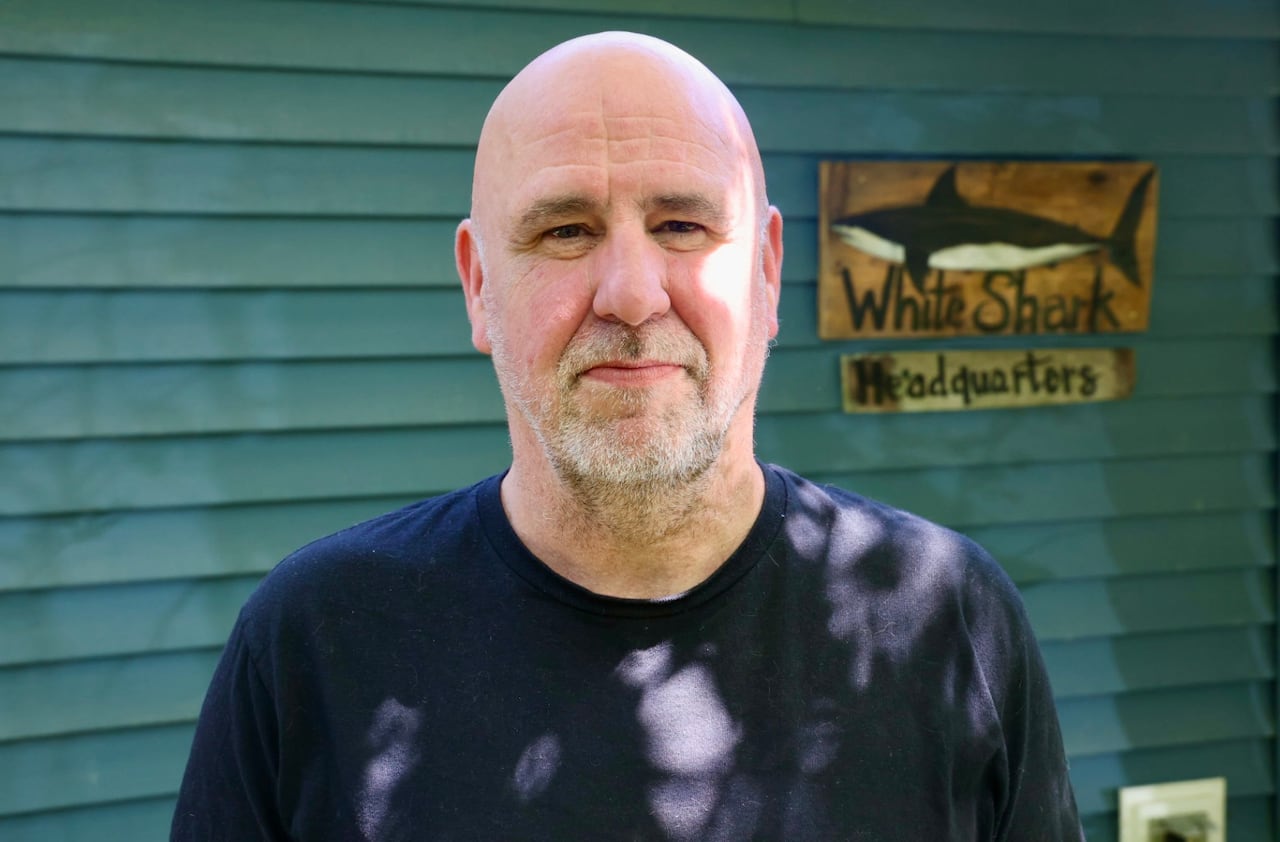 A bald man with a navy blue shirt stands in front of blue siding. A sign can be seen behind him that reads "White Shark Headquarters"