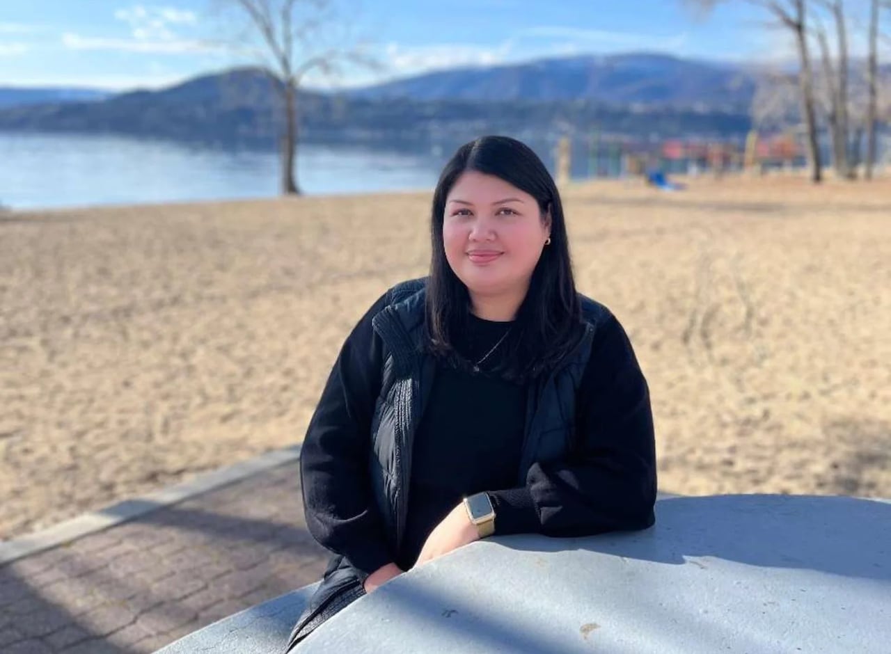 A smiling woman with long dark hair sits at a table outdoors, a beach seen behind her.