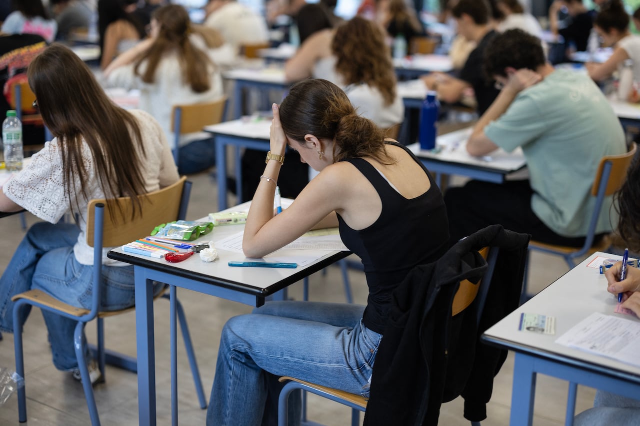A student in a blank tank top and jeans rests her head in one hand as she sits to write an exam, with fellow students also writing the exam seen around her.