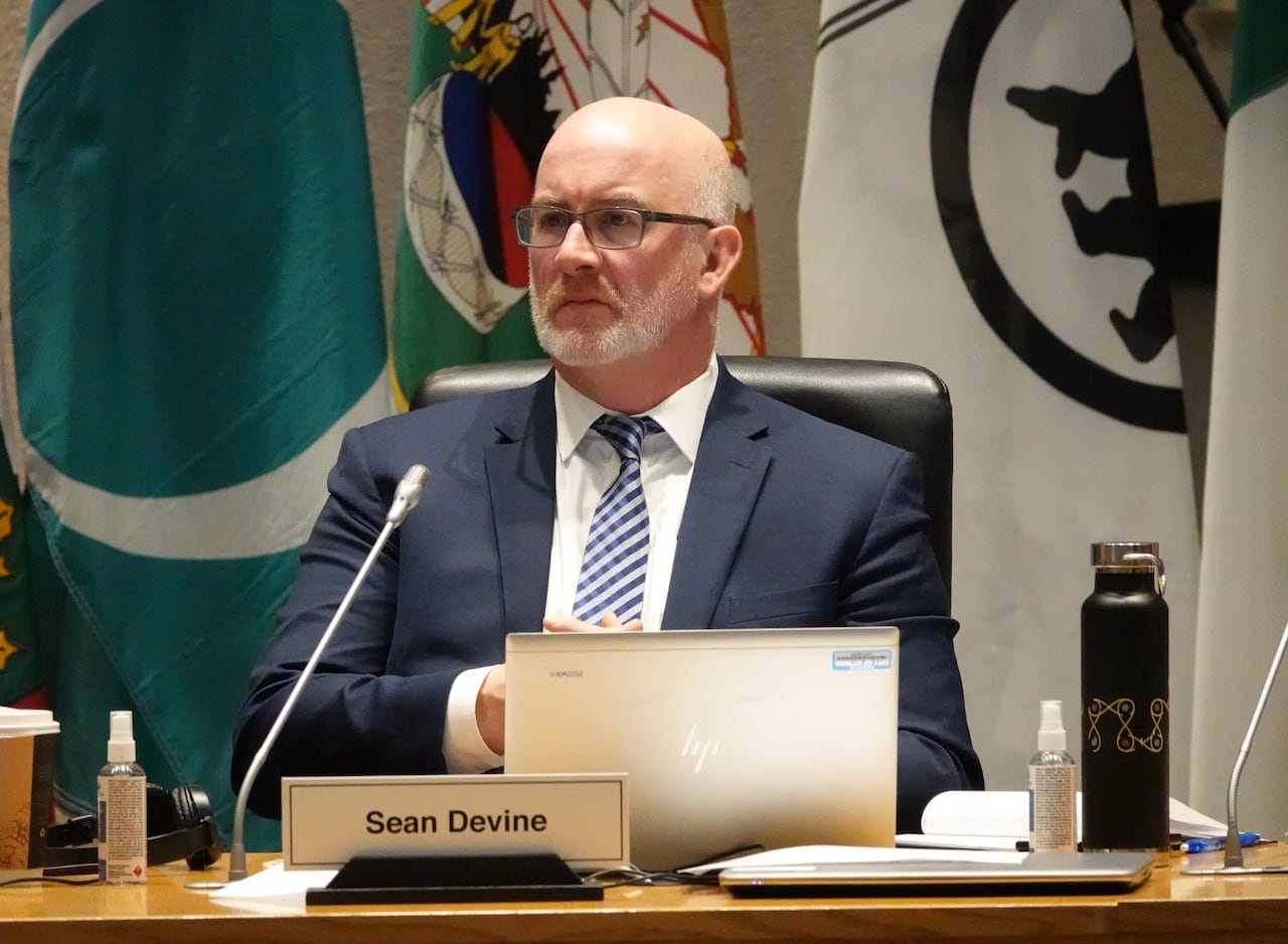 A city councillor sits at a desk with their nameplate on it, listening during a meeting.