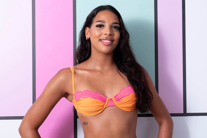 Love Island S13 Nevaeh stands with her hands on her hips, smiling and is wearing a bright orange bikini with pink trim. She has dark brown hair.