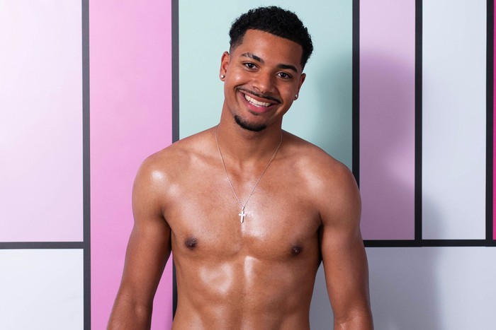 Love Island S13 Carlos stands in front of a colourful backdrop and is topless. He smiles and has dark brown hair.