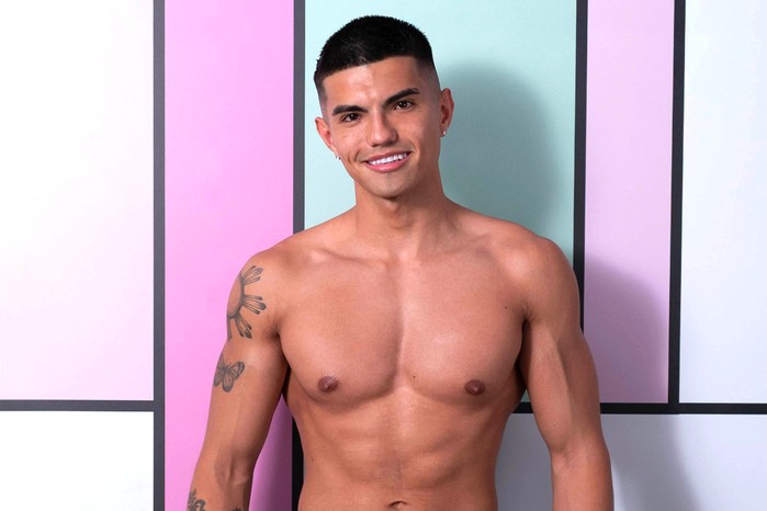 Love Island S13 Will stands in front of a colourful background and is topless. He has tattoos and is smiling.