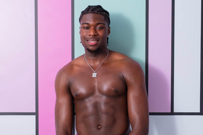 Love Island S13 Aaron stands in front of a colourful backdrop and is topless. He has dark brown hair which is plaited backwards and is wearing a necklace.