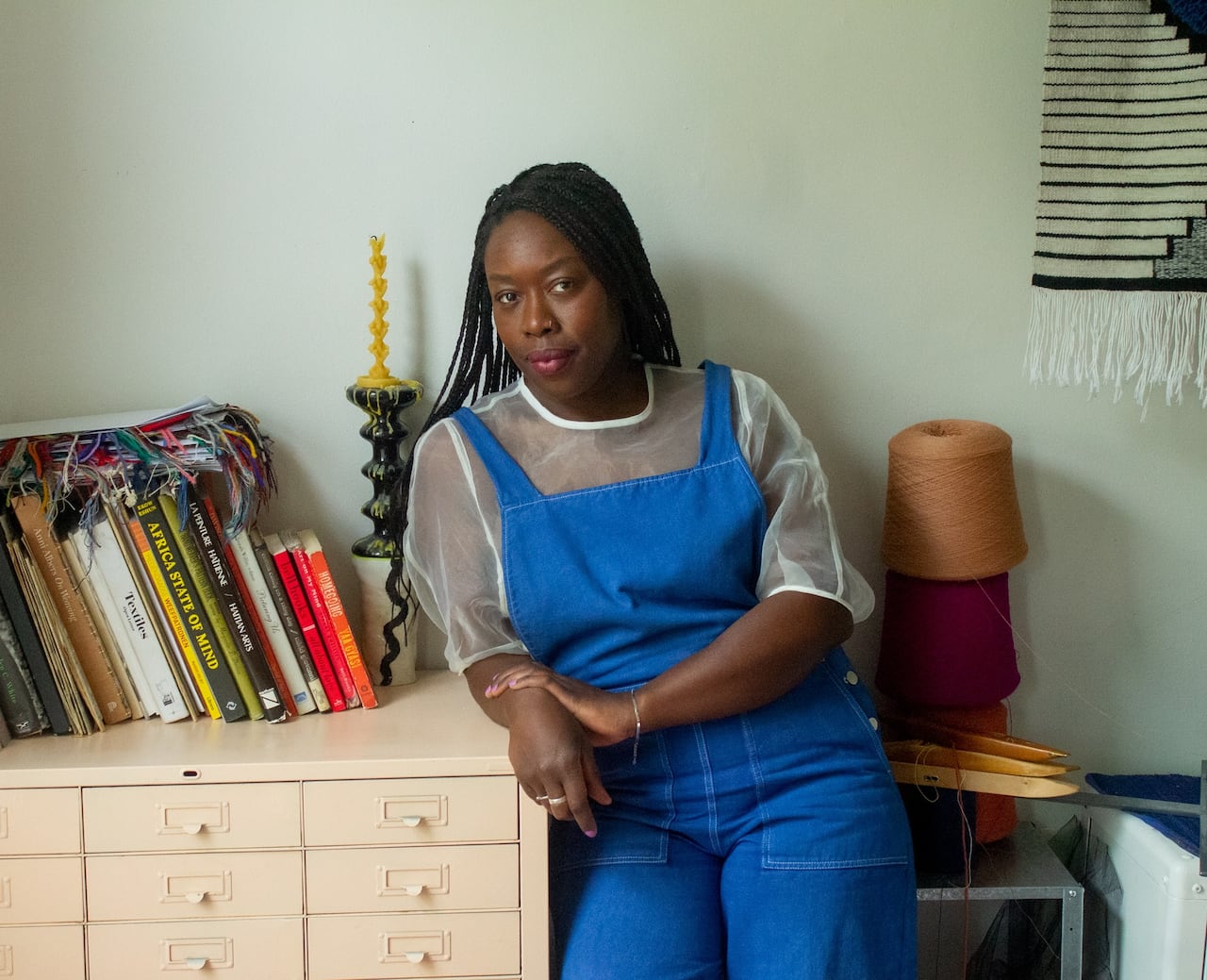 A Black woman poses in blue overalls next to the textiles she uses in her artwork.