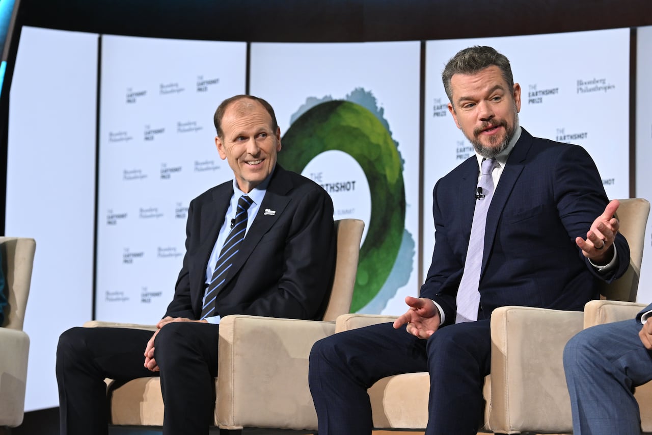 Two men sit in beige armchairs on a stage during a presentation. The man on the left is balding and wearing a navy suit, light blue shirt and a blue striped tie. A bearded man on the right is in a navy suit, white button down shirt and mauve necktie.