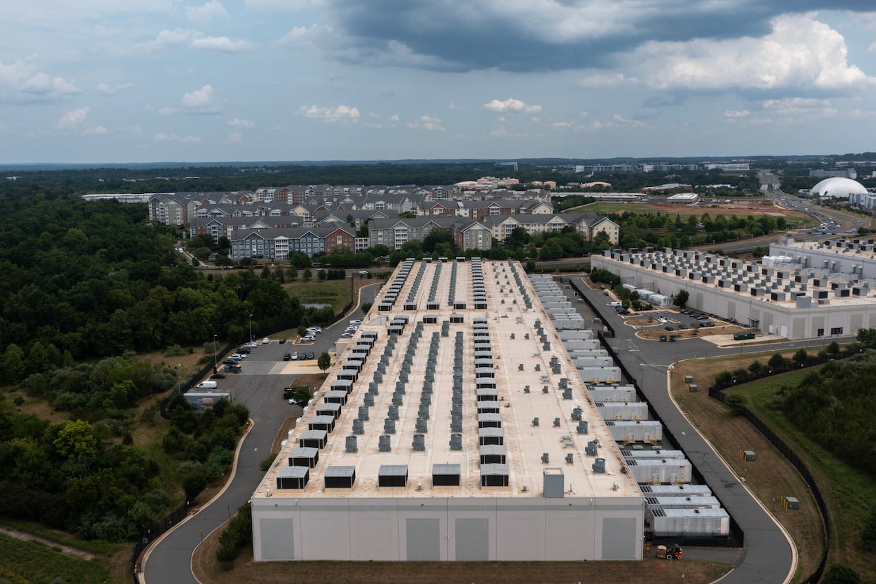 A drone camera view of a long concrete building housing a data centre, with cooling units on the roof. Two similar structures are seen to the right and rows of residential buildings are situated in the background.