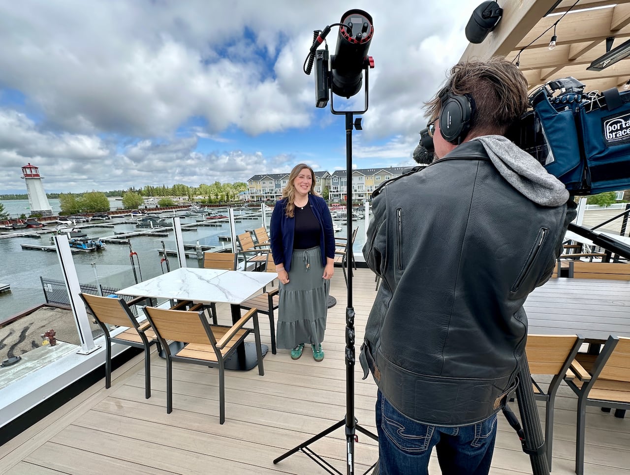 A woman in a long skirt, standing on a patio with the harbour the background talking to a a camera man.