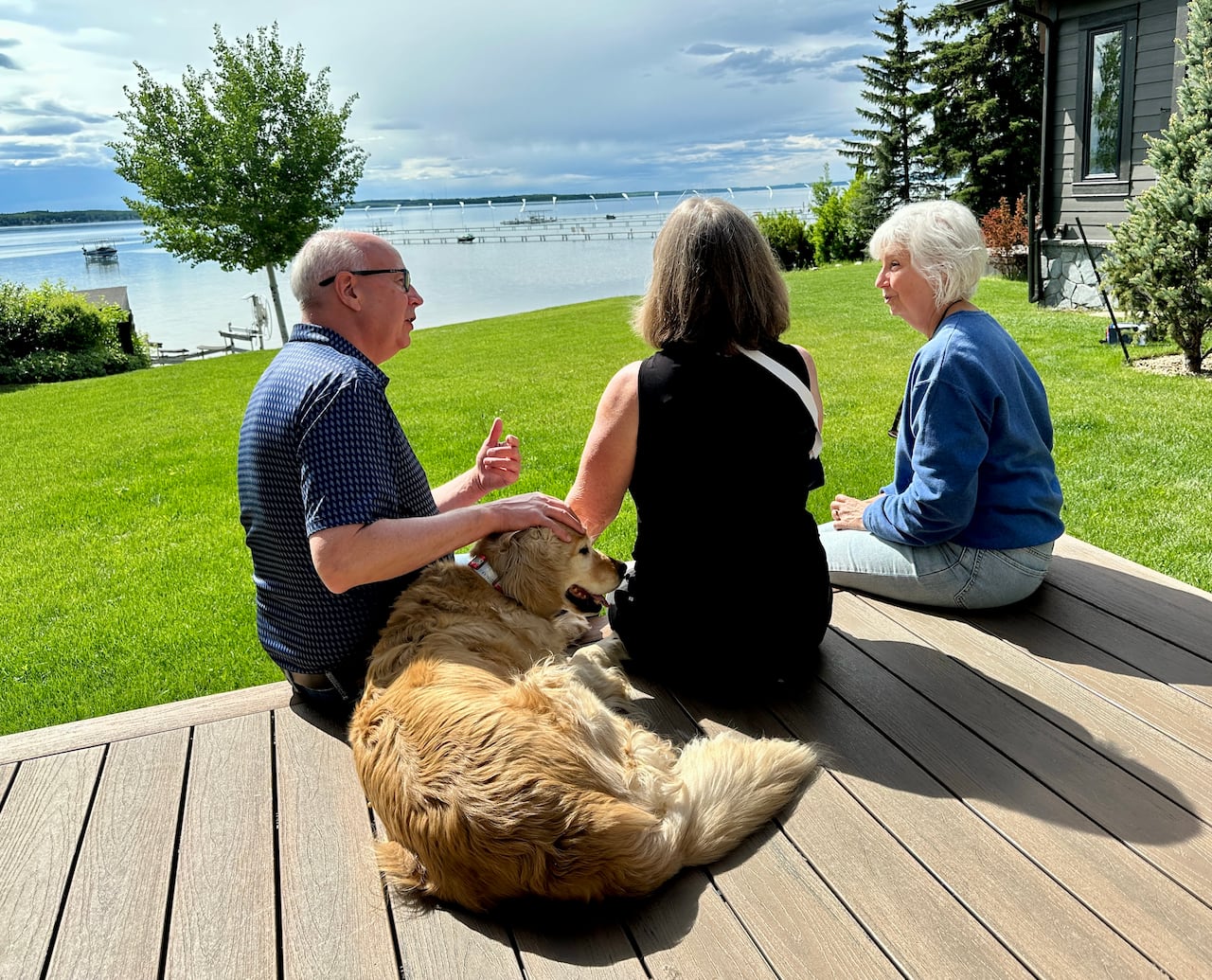 Three people sit next to a pretty large golden dog with the lake and trees in the background