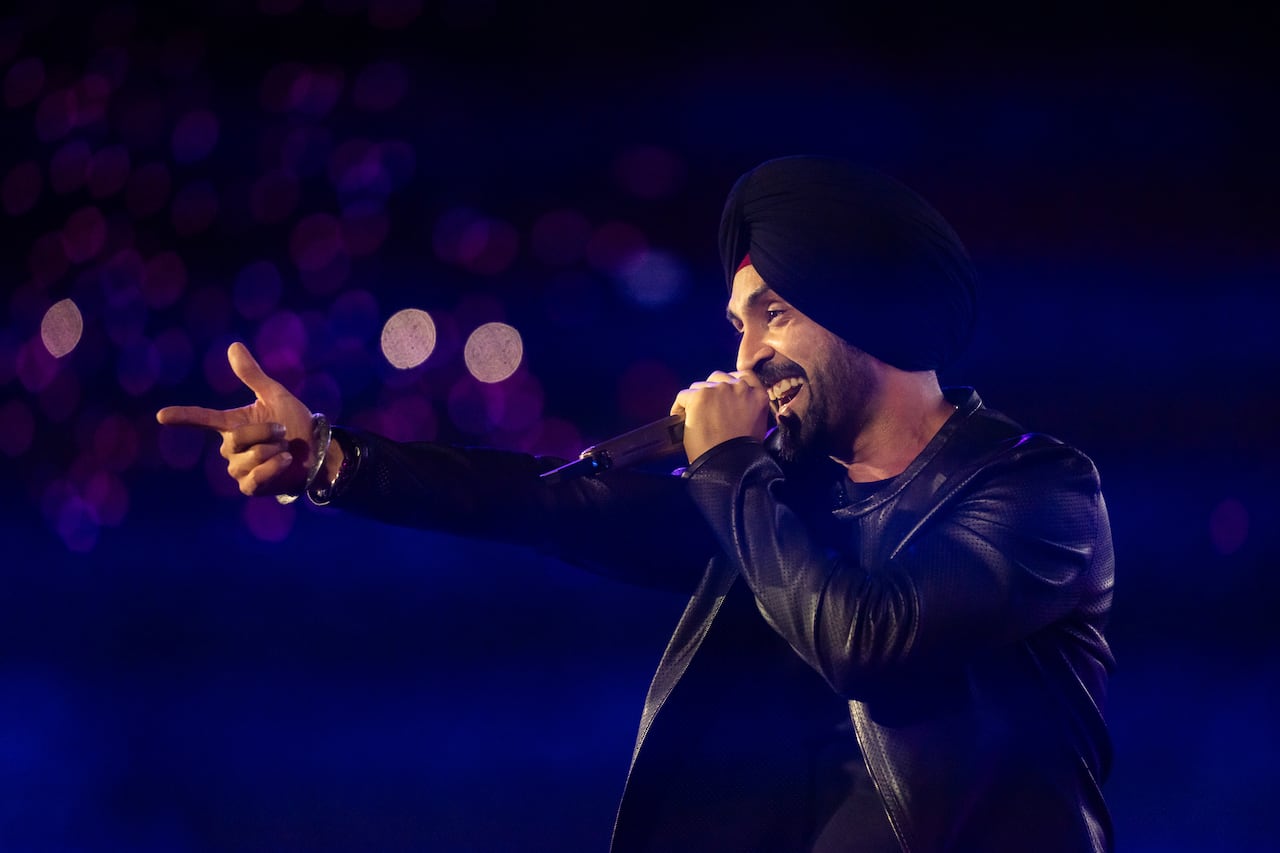 A Sikh man wearing black turban, T-shirt and leather jacket holding a mic and performing on stage.