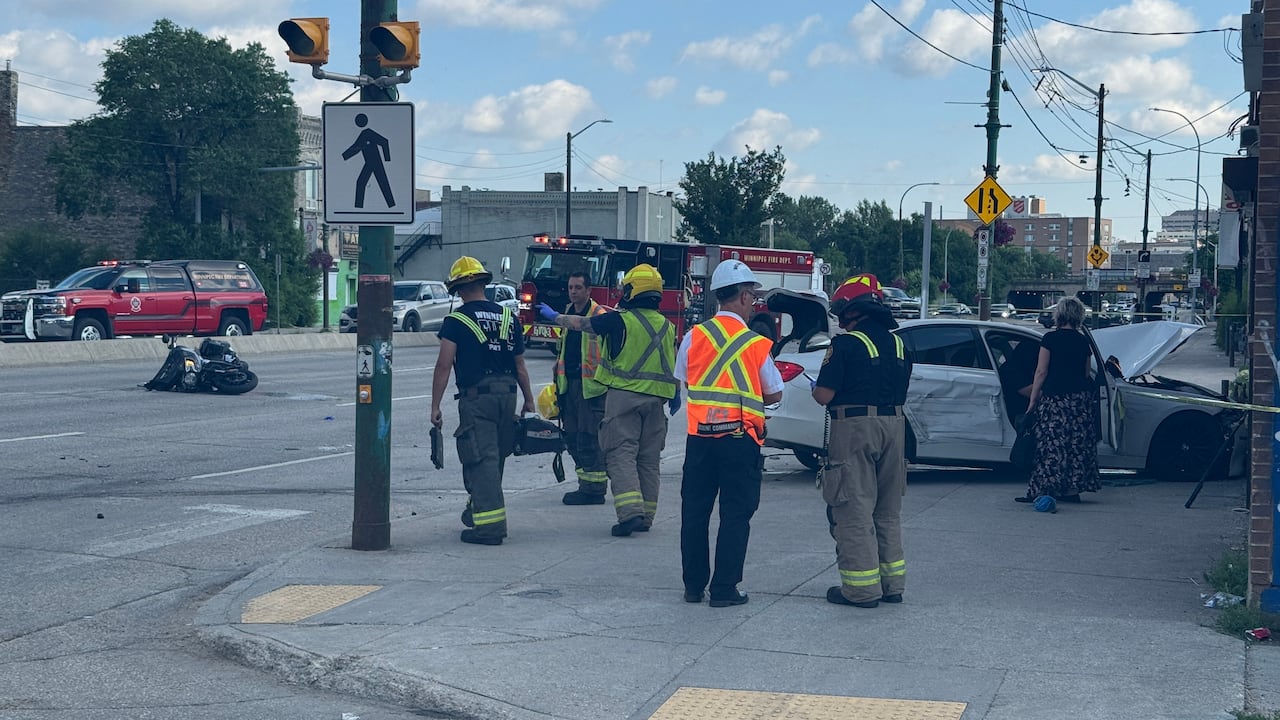 Firefighters stand at the side of a road, where a motorcycle lies crumpled. A silver sedan is crashed into the sidewalk